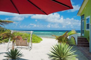 Un sentier bordant la plage mène à une maison surmontée d’un parasol rouge, entourée de plantes tropicales et offrant une vue sur l’océan.