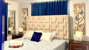 Ein Schlafzimmer mit einer gesteppten Kopfteil, einem Bett mit einem gemusterten Bettbezug, einem blauen Vorhang, einem Nachttisch mit Lampe und einem wandmontierten Spiegel.
