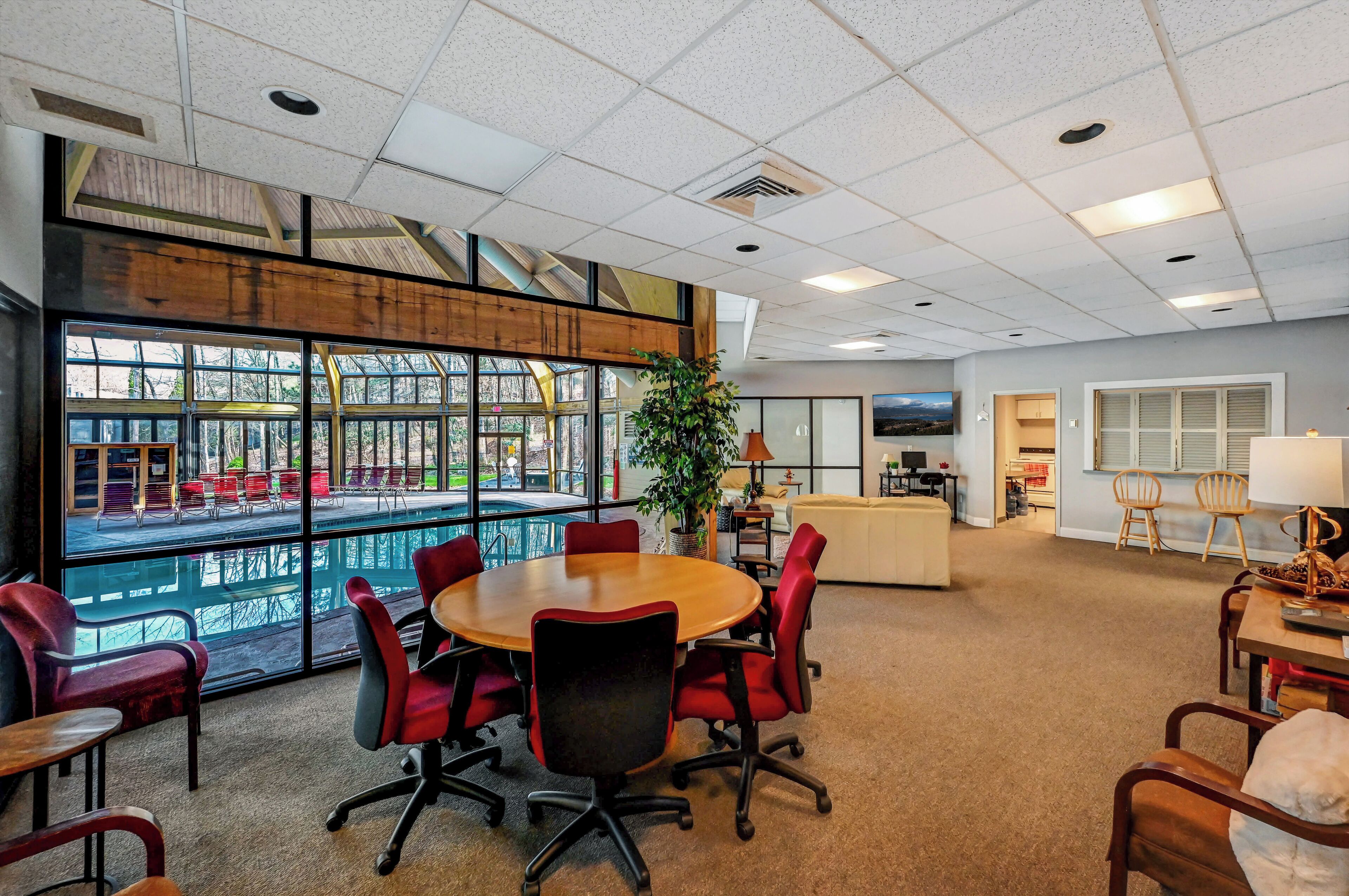 A conference room with a large table and red chairs, large windows with a view of outdoor seating, and a plant in the center.