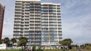 Ein mehrstöckiges Hotel mit Balkonen, einem zentralen, gelben vertikalen Element und einer Strandlage.