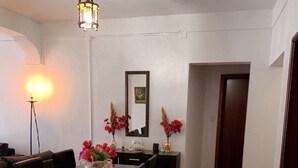 Un coin repas avec une table dressée pour quatre personnes, entourée de chaises. On y voit une lampe sur pied et un miroir au mur. Une plante est visible dans un coin.