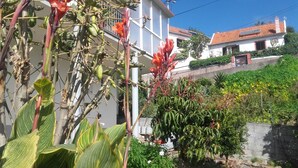 Un immeuble résidentiel à la façade blanche et au toit recouvert de tuiles rouges, entouré d’une végétation luxuriante et de plantes en fleurs.