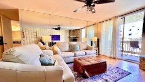 A spacious living room with a large sofa, a coffee table, and a TV. There is a ceiling fan and a balcony with a chair. - OCEANFRONT White Water Views Unit E-100  SEASONAL RATES AVAILABLE PLEASE INQUIRE (Oceanside)