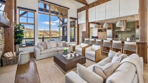Un salón amplio con ventanas grandes, una zona de estar acogedora y una cocina moderna visible a través de una puerta abierta.