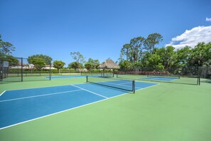 Una cancha de tenis con red, rodeada de árboles y una estructura con techo de paja.