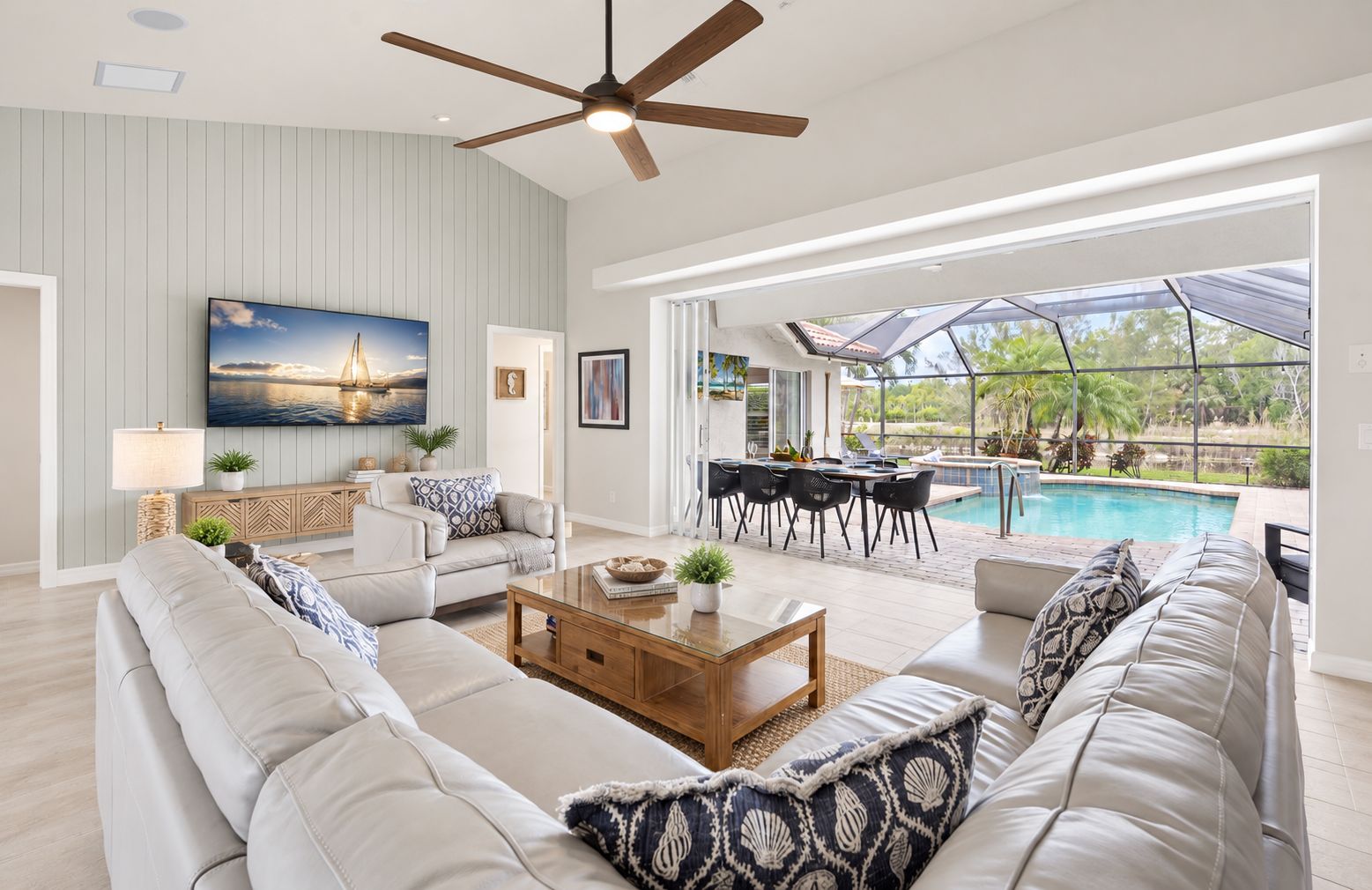 A spacious living room with a large sofa, a coffee table, and a ceiling fan. There is a dining area with a table and chairs, and a view of a swimming pool.