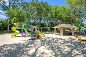 Un parque infantil con toboganes y estructuras para escalar, rodeado de árboles y una zona de estar cubierta.