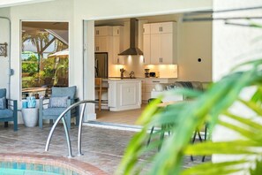 Una cocina moderna con armarios blancos y electrodomésticos de acero inoxidable, un comedor con mesa y sillas, y un área de estar junto a la piscina con vistas al exterior.