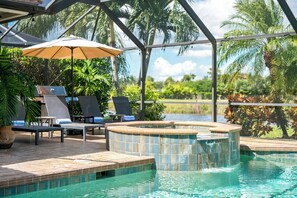 Zona de piscina con sillones reclinables, jacuzzi y vistas a un lago rodeado de palmeras.