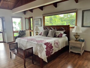 A bedroom with a wooden bed, a nightstand with a lamp, a chair, and a window with a view of greenery. - VERY PRIVATE, expansive ocean VIEWS, close to BEST BEACH (Kailua-Kona)