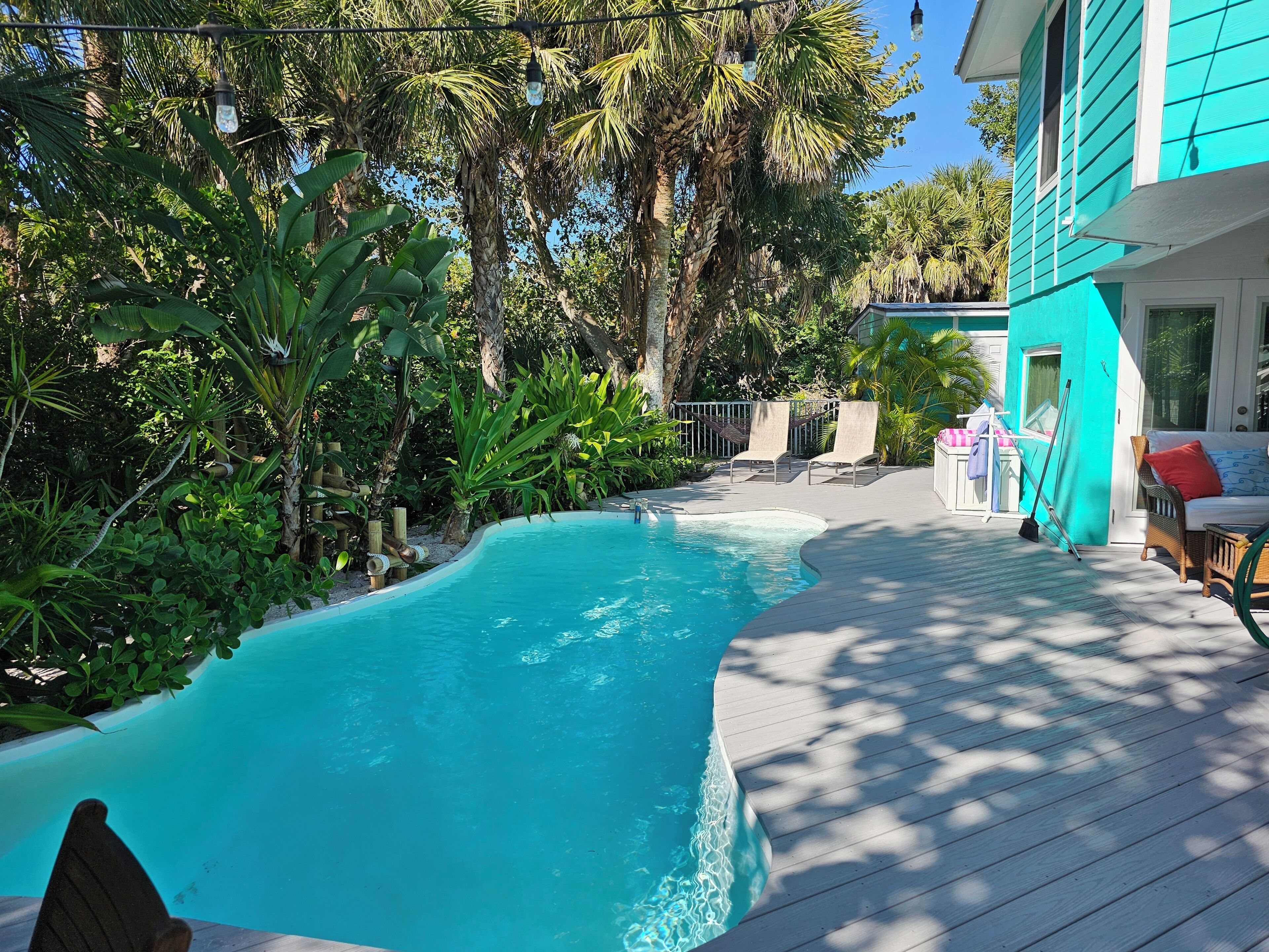 A swimming pool surrounded by lush greenery, a wooden deck, and a turquoise building.