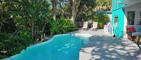 A swimming pool surrounded by lush greenery, a wooden deck, and a turquoise building.