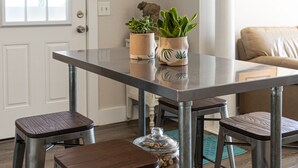 Un coin repas comprenant une table à plateau en verre, quatre tabourets de bar en métal et deux plantes en pot.