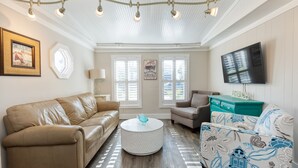 Un salon avec un canapé beige, un fauteuil à motifs, une table basse ronde et un téléviseur à écran plat fixé au mur.