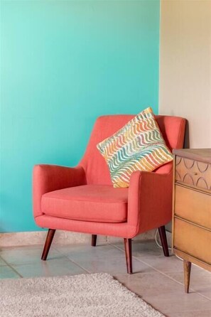 Un fauteuil rouge avec un coussin à motifs, adossé à un mur de couleur turquoise, une commode en bois et un tapis blanc sur un sol carrelé.