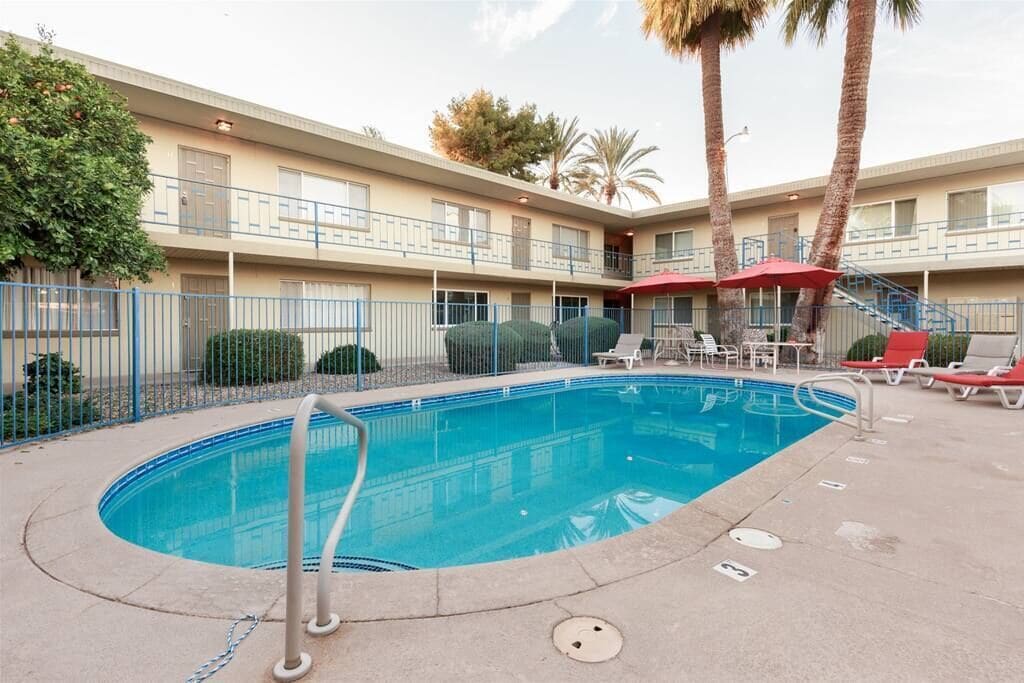 A swimming pool with a blue deck, surrounded by a fence, and a building with balconies and palm trees in the background.