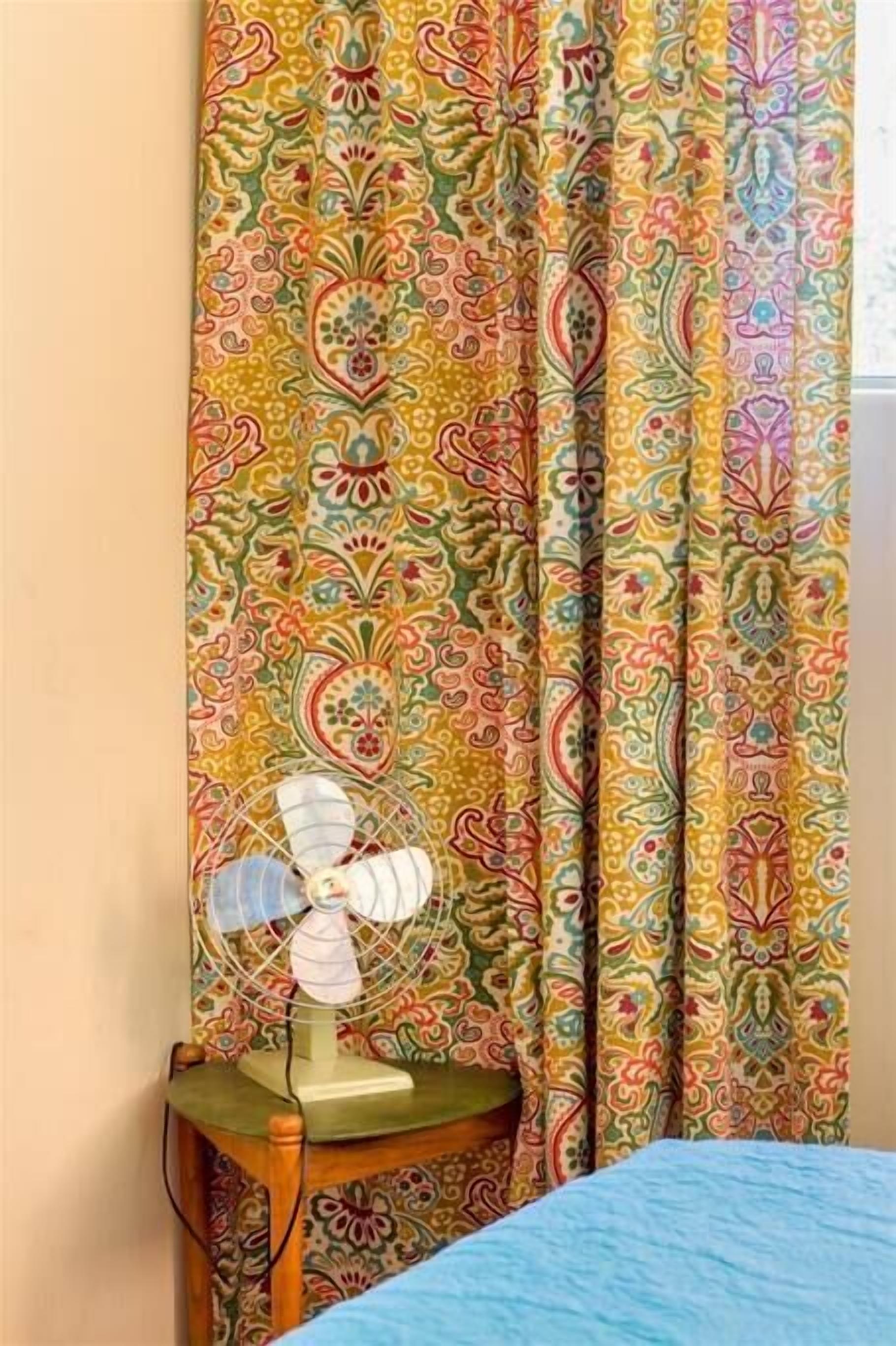Un letto con copriletto blu, un comodino in legno con una ventola bianca e una tenda a motivi paisley.
