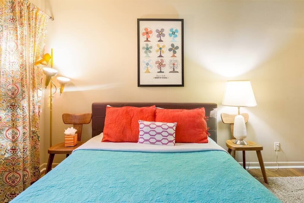 Een net opgemaakt bed met een blauwe sprei, twee oranje kussens en een lijst met foto's van supporters aan de muur.