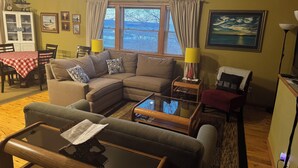 A living room with a sectional sofa, a glass coffee table, a dining table with a red and white checkered tablecloth, and a painting on the wall. - Private And Secluded Rental On the Kalamazoo River - Saugatuck-Douglas-Fennville (Fennville)