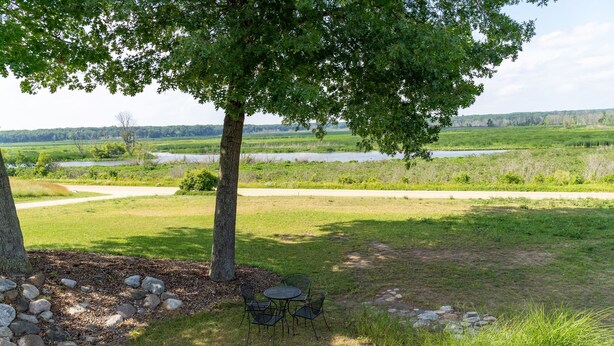 A grassy outdoor area with a table and chairs under a tree, surrounded by rocks and a view of a lake and fields. - Private And Secluded Rental On the Kalamazoo River - Saugatuck-Douglas-Fennville (Fennville)