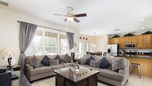 A modern living room with a glass coffee table, two sofas, a ceiling fan, and a kitchen area with wooden cabinets and appliances. - 🏰 Magical 6-Bed Family Villa ✨[FREE Heated Pool, Spa & BBQ] 5 Mins to Disney 🎢 (Kissimmee)