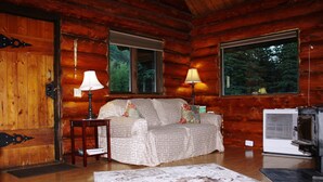 Een knus interieur van een blokhut met een houten wand, een bank met gestreepte kussens, twee lampen en een groot raam met uitzicht op het bos.