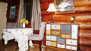 Een houten blokhutinterieur met een eettafel, een tafelkleed met bloemen, een lamp, een dressoir met lades, gordijnen en een lijst met een landschapsafbeelding.