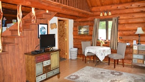 Een houten blokhut van binnen, met een houten trap, een televisie op een kast, een eettafel met stoelen, en een raam met gordijnen.