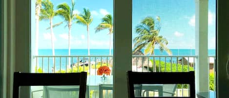 A room with two wooden chairs facing a balcony with a view of the ocean and palm trees.