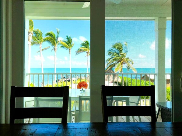 A room with two wooden chairs facing a balcony with a view of the ocean and palm trees.