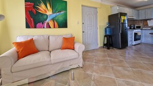 Un soggiorno con un divano beige, due cuscini arancioni, un quadro colorato appeso al muro, una ventola a soffitto e un angolo cottura con frigorifero, microonde e fornello.