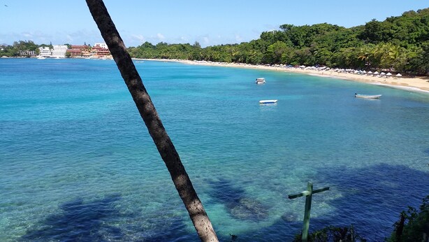 Una spiaggia con acque turchesi cristalline, alcune barche e una croce sullo sfondo.