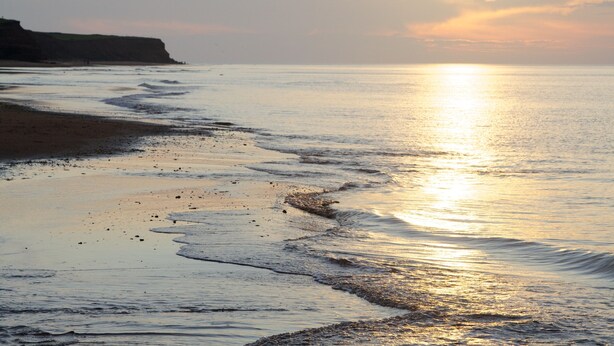 A beach with gentle waves, a clear sky, and a distant cliff.