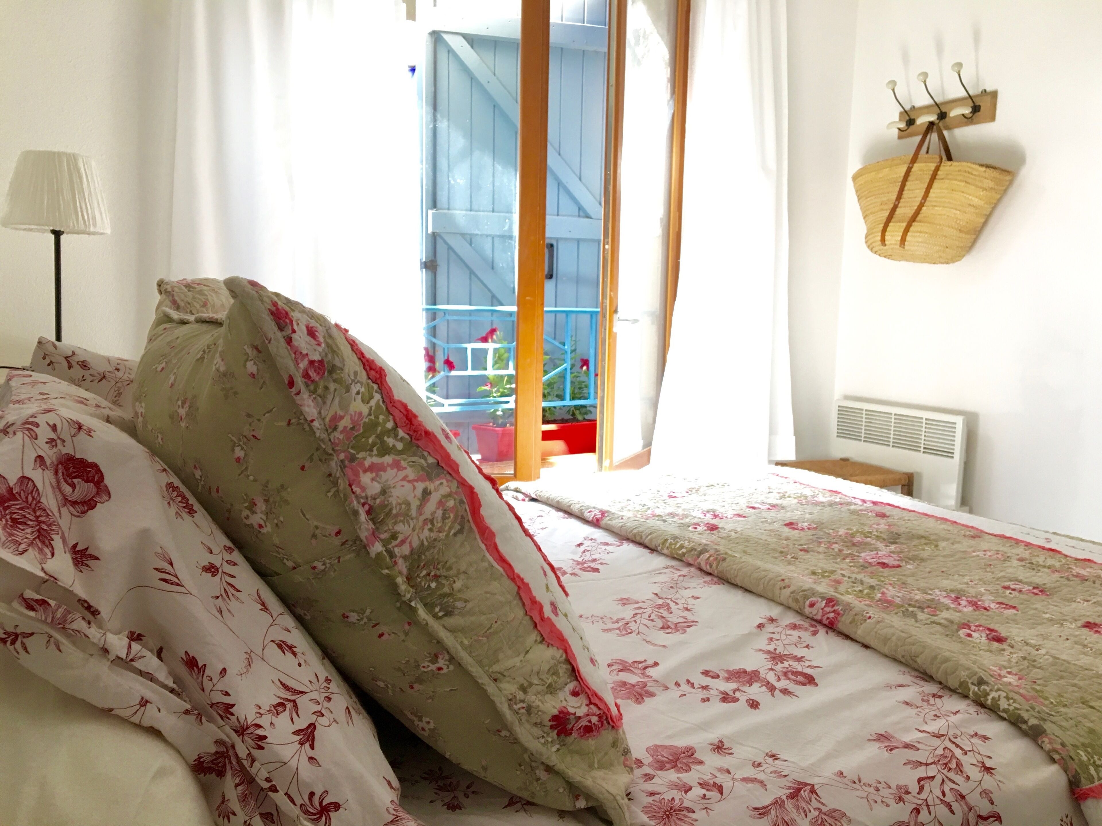 A bedroom with floral bedding, a bedside lamp, a window with curtains, and a wall-mounted hook with a bag.