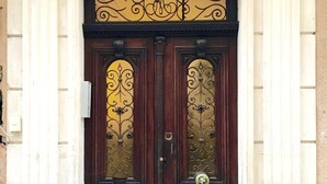 Une porte en bois à deux battants, ornée de motifs métalliques complexes, flanquée de colonnes blanches et surmontée d’une décoration sculptée.