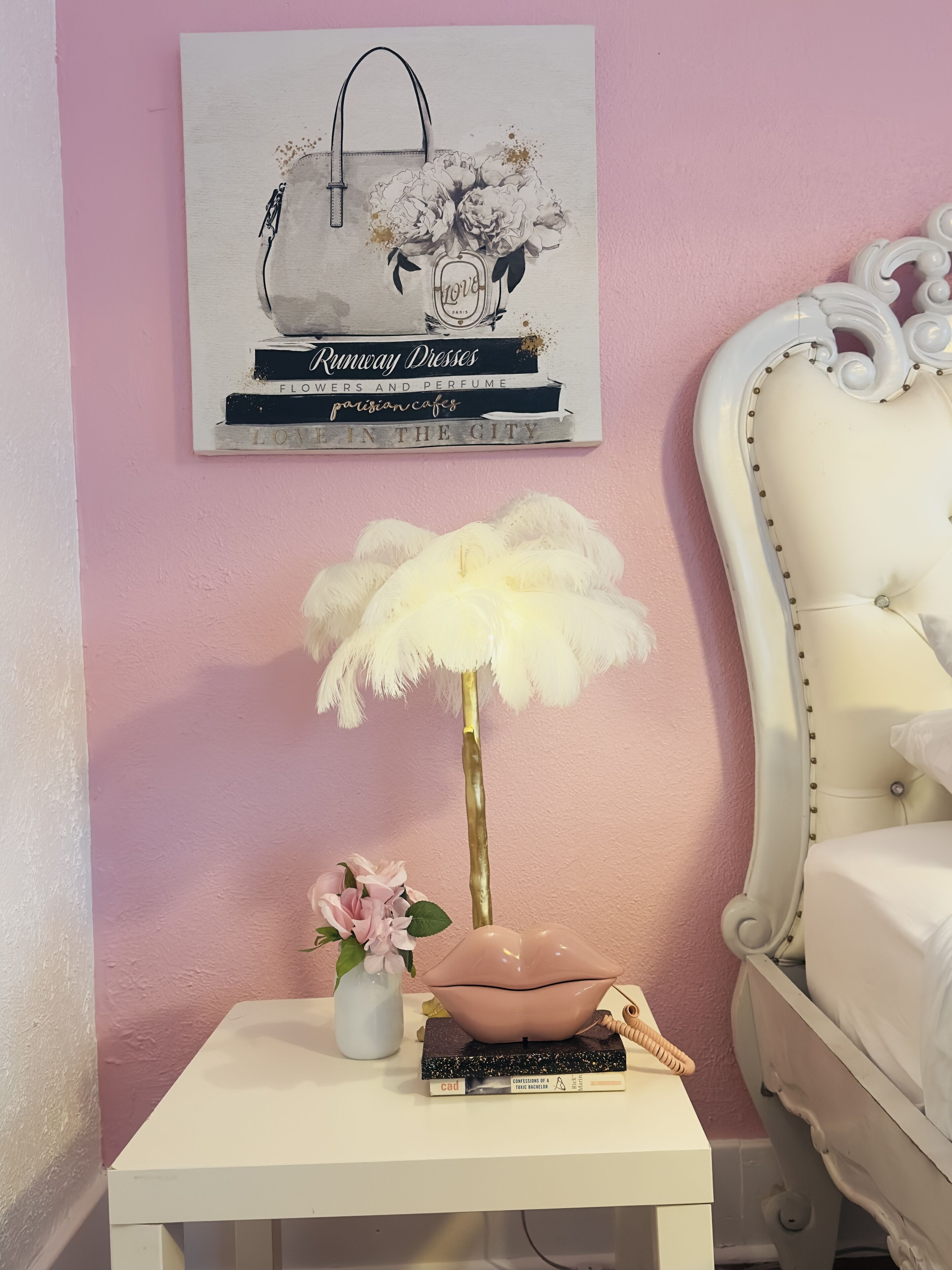 A neatly arranged bedside table with a vase of pink flowers, a decorative bowl, and a book titled "Conversations of a..." against a pink wall.