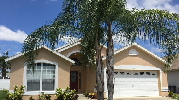 A single-story house with a beige exterior, a white garage door, and a well-maintained lawn with a palm tree. - Pet-Friendly Fenced Home w/Pool & Spa Heat - 3 Miles from Disney (Kissimmee)