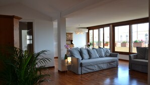 Uma sala de estar ampla com um sofá cinza, piso de madeira, janelas grandes e uma planta em vaso.