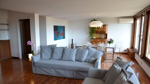 Uma sala de estar moderna com um sofá cinza, piso de madeira, área de jantar com mesa e cadeiras, e cozinha ao fundo.