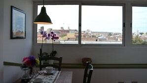 Uma sala de jantar com uma mesa posta para dois, um vaso com flores e vista para a cidade através de grandes janelas.