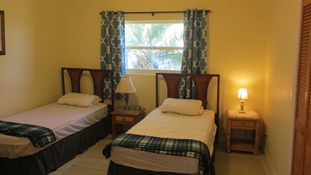 A room with two single beds, each with a bedside table and lamp, a window with patterned curtains, and a wooden ceiling. - LARGE 4 Bed/4Bath BEACHFRONT APARTMENT SLEEPS 8 and directly  on HOOPERS BAY (Georgetown)