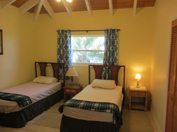 A room with two single beds, each with a bedside table and lamp, a window with patterned curtains, and a wooden ceiling. - LARGE 4 Bed/4Bath BEACHFRONT APARTMENT SLEEPS 8 and directly  on HOOPERS BAY (Georgetown)