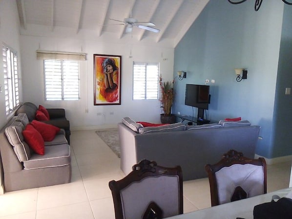 A spacious living area with a grey sofa, red cushions, a large painting, a flat-screen TV, and a ceiling fan.