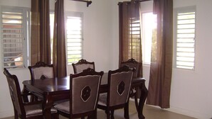 A dining area with a wooden table and chairs, curtains, and windows with blinds. - OCHO RIOS Jamaica Paradise House 2 (St Ann's Bay)