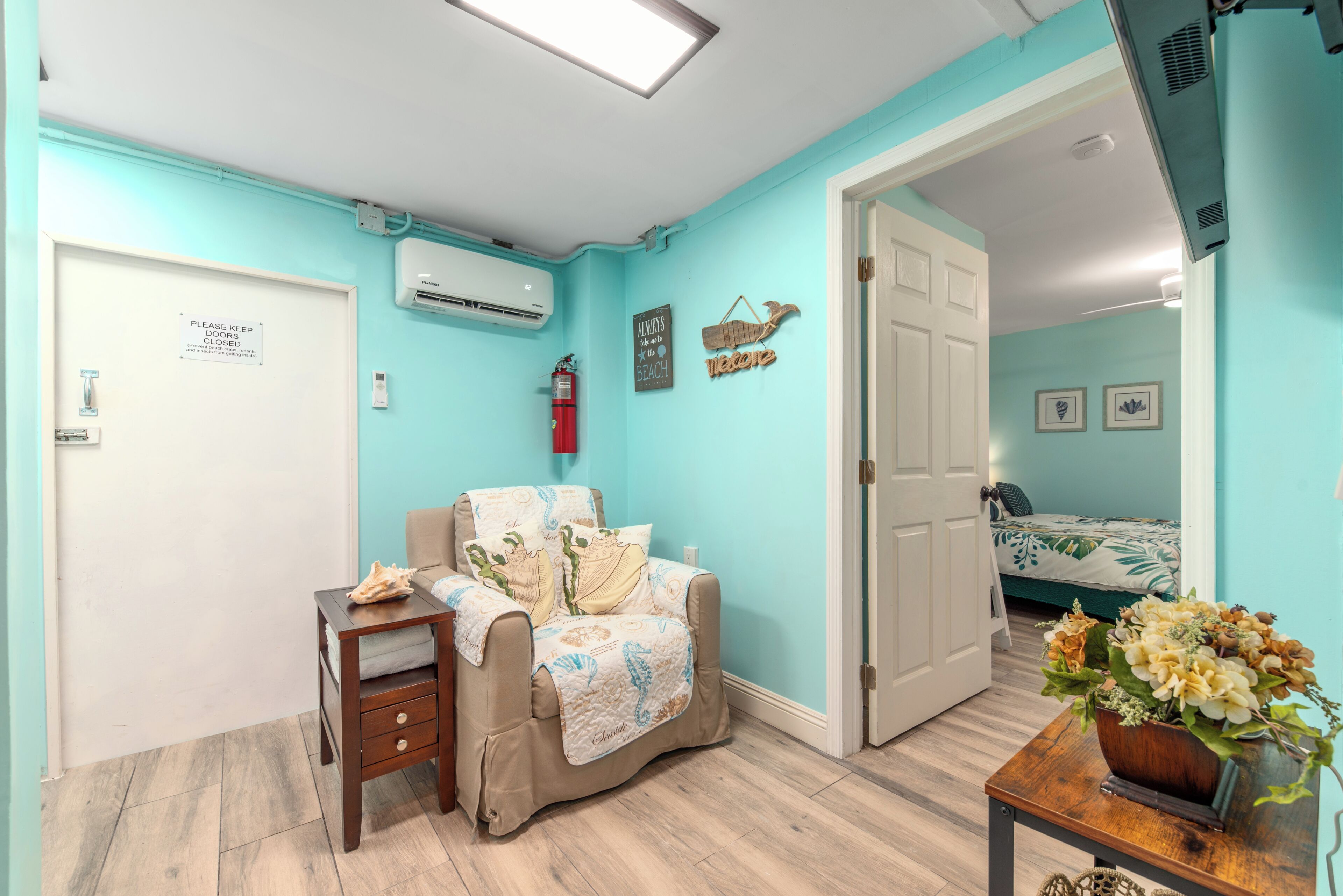 A room with teal walls, a bed with a floral bedspread, a chair with a blanket, a small table with a vase, and a door with a sign.