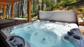 Un bain à remous doté d’un panneau de commande, entouré d’une terrasse en bois et bordé d’une colline boisée.