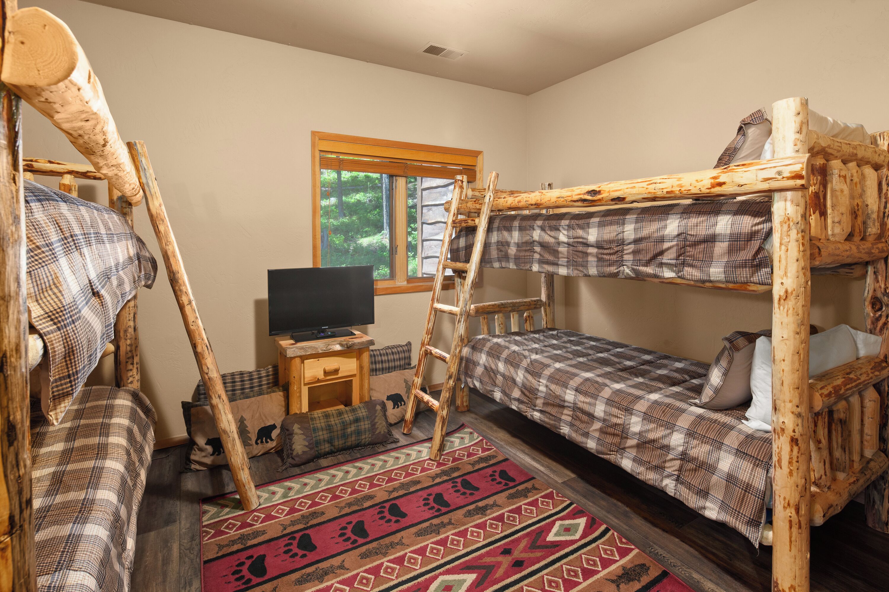 A rustic room with two bunk beds, a television on a wooden stand, a window with a view of trees, and a patterned rug.