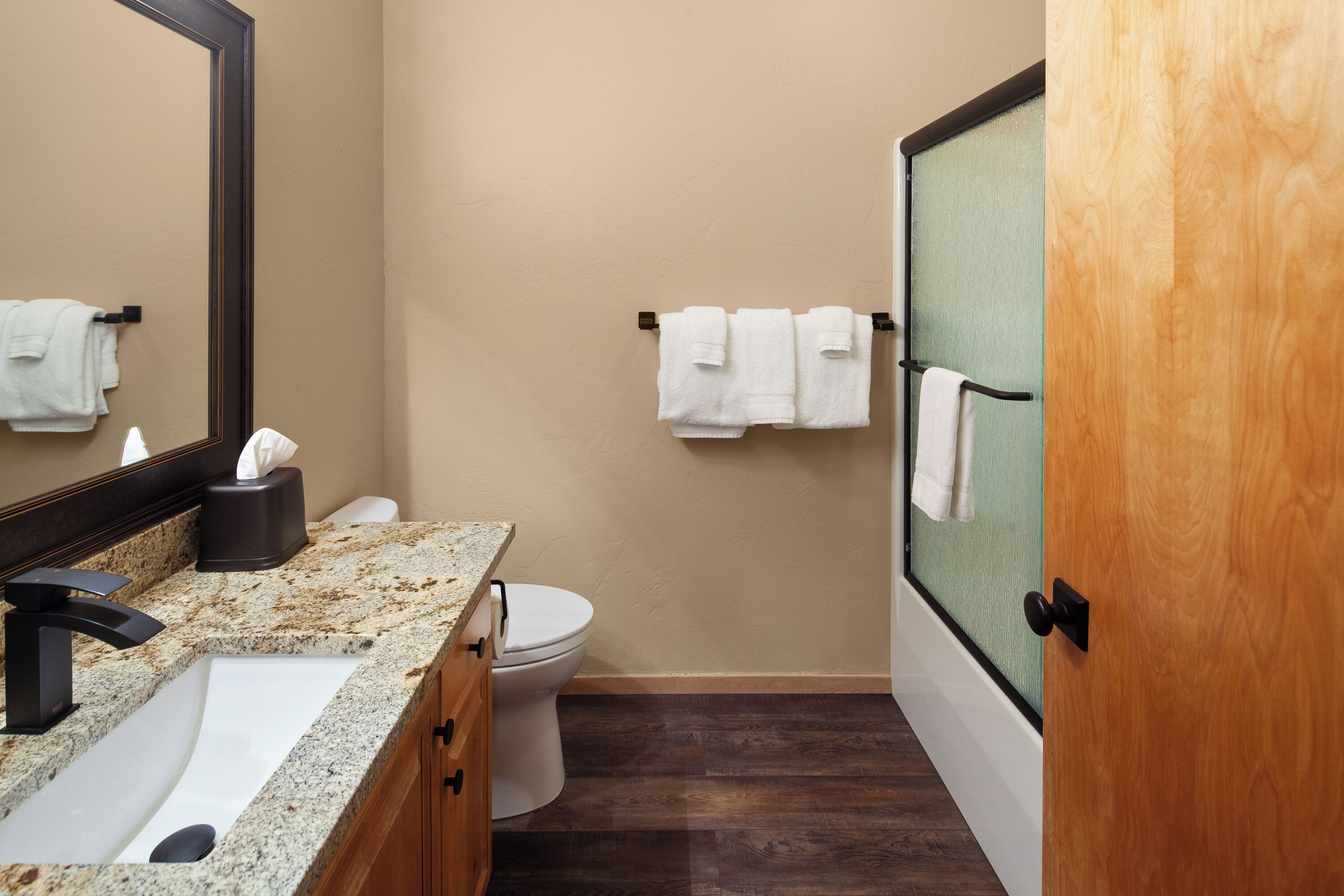 A bathroom with a granite countertop, a black faucet, a white sink, a toilet, and a wooden door.