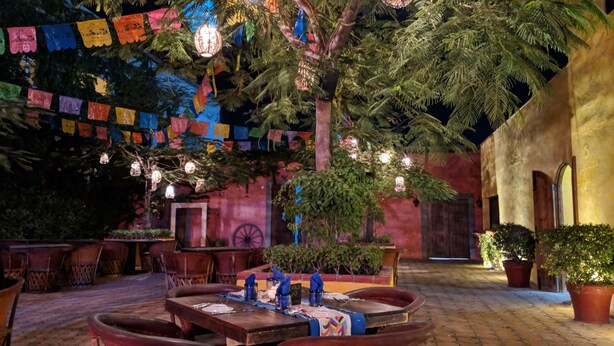 Een terras met tafels voor twee personen, omgeven door potplanten en een landelijk gebouw op de achtergrond.