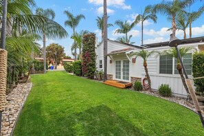 A well-maintained lawn with a wooden deck, palm trees, and a white building with a green door. - Plantation Beach Cottage - Historic Encinitas Walking District (Encinitas)